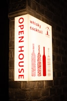 An illuminated sign is mounted on a brick wall, displaying the words 'OPEN HOUSE' vertically in bold red letters. To the right, it features the words 'whisky cocktail' and stylized images of wine bottles, also in red. The background is a soft white, creating a striking contrast with the red text and images.