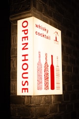 An illuminated sign is mounted on a brick wall, displaying the words 'OPEN HOUSE' vertically in bold red letters. To the right, it features the words 'whisky cocktail' and stylized images of wine bottles, also in red. The background is a soft white, creating a striking contrast with the red text and images.