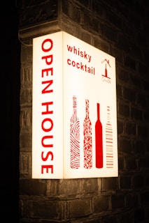 An illuminated sign is mounted on a brick wall, displaying the words 'OPEN HOUSE' vertically in bold red letters. To the right, it features the words 'whisky cocktail' and stylized images of wine bottles, also in red. The background is a soft white, creating a striking contrast with the red text and images.