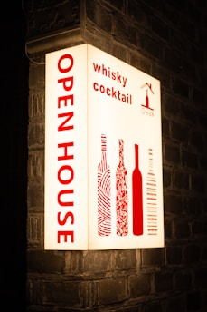 An illuminated sign is mounted on a brick wall, displaying the words 'OPEN HOUSE' vertically in bold red letters. To the right, it features the words 'whisky cocktail' and stylized images of wine bottles, also in red. The background is a soft white, creating a striking contrast with the red text and images.