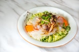 A beautifully arranged dish of cold noodles topped with fresh vegetables and herbs. There are sliced cucumbers, carrots, and radishes arranged around the edge of a white plate, with translucent noodles and a garnish of cilantro and chili flakes in the center.