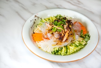 A beautifully arranged dish of cold noodles topped with fresh vegetables and herbs. There are sliced cucumbers, carrots, and radishes arranged around the edge of a white plate, with translucent noodles and a garnish of cilantro and chili flakes in the center.