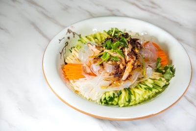 A beautifully arranged dish of cold noodles topped with fresh vegetables and herbs. There are sliced cucumbers, carrots, and radishes arranged around the edge of a white plate, with translucent noodles and a garnish of cilantro and chili flakes in the center.