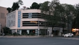 A multi-story building with a modern design, featuring large windows and a prominent vertical sign with Chinese characters. The building is situated on a street corner, surrounded by trees and some greenery on the roof. The area appears to be quiet, with minimal traffic, and a few cars are visible on the road.