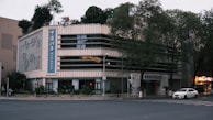 A multi-story building with a modern design, featuring large windows and a prominent vertical sign with Chinese characters. The building is situated on a street corner, surrounded by trees and some greenery on the roof. The area appears to be quiet, with minimal traffic, and a few cars are visible on the road.