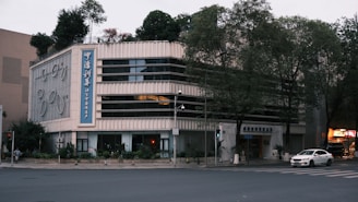 A multi-story building with a modern design, featuring large windows and a prominent vertical sign with Chinese characters. The building is situated on a street corner, surrounded by trees and some greenery on the roof. The area appears to be quiet, with minimal traffic, and a few cars are visible on the road.