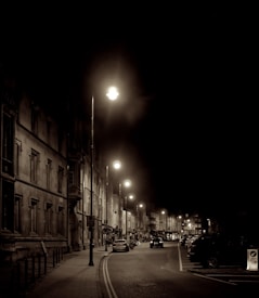 A nighttime urban street scene with vintage-style sepia tones. Streetlights line the road, casting a warm glow on the surrounding buildings and sidewalk. Several cars are parked along the side, and a few vehicles are driving through the scene.