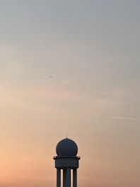 A radar tower with a spherical dome on top stands against a backdrop of a serene, color-gradient sky transitioning from light blue at the top to shades of orange and pink near the horizon. An airplane is visible in the distance, adding a sense of scale and activity to the scene.