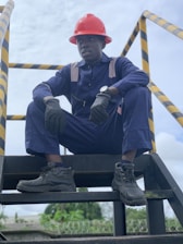 A person wearing a navy blue work uniform and a red hard hat is sitting on an industrial stairway. The individual is also wearing black gloves and black work boots. Yellow and black caution striped handrails are visible on either side.