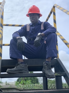 A person wearing a navy blue work uniform and a red hard hat is sitting on an industrial stairway. The individual is also wearing black gloves and black work boots. Yellow and black caution striped handrails are visible on either side.
