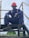 A person wearing a navy blue work uniform and a red hard hat is sitting on an industrial stairway. The individual is also wearing black gloves and black work boots. Yellow and black caution striped handrails are visible on either side.