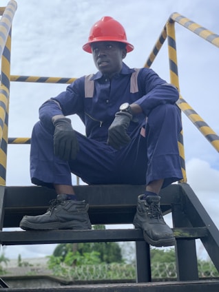 A person wearing a navy blue work uniform and a red hard hat is sitting on an industrial stairway. The individual is also wearing black gloves and black work boots. Yellow and black caution striped handrails are visible on either side.