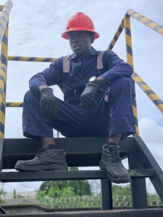 A person wearing a navy blue work uniform and a red hard hat is sitting on an industrial stairway. The individual is also wearing black gloves and black work boots. Yellow and black caution striped handrails are visible on either side.