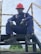 A person wearing a navy blue work uniform and a red hard hat is sitting on an industrial stairway. The individual is also wearing black gloves and black work boots. Yellow and black caution striped handrails are visible on either side.