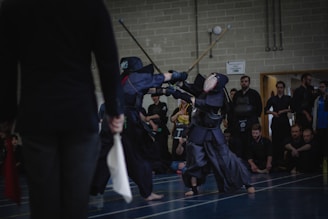 Two individuals engaged in a kendo match, dressed in traditional protective armor and wielding bamboo swords, are captured mid-action on a sports court. A referee stands nearby holding flags, while a group of spectators watches the match intently from the sidelines.
