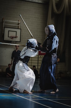 Two individuals are engaged in a kendo match, wearing traditional protective gear and wielding shinai sticks. The scene is set indoors on a sports court with a basketball hoop visible in the background.