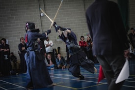 Two individuals are engaged in a Kendo match, wearing traditional protective gear and wielding bamboo swords. They are observed by a small audience in a gymnasium setting, with other spectators seated and standing in the background.