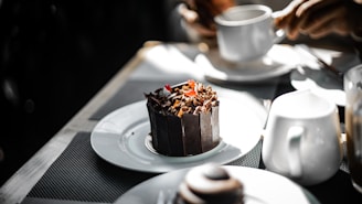 A dark chocolate dessert garnished with chocolate shavings and a small red fruit is placed on a white plate. Nearby, a person holds a white cup, suggesting a setting of dining or having a coffee. Other white crockery items are present on the table, which is covered with a textured, dark-colored mat, creating a contrasting background for the dessert.