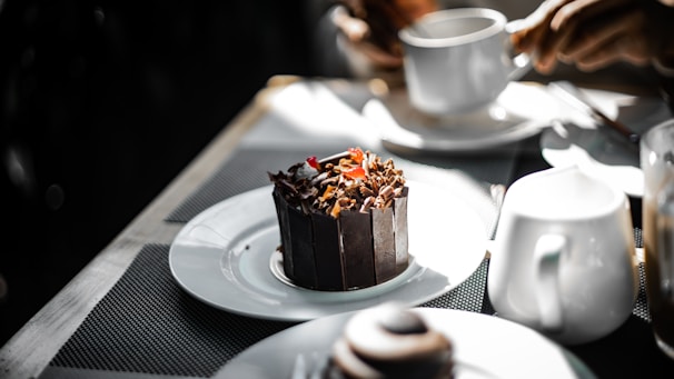 A dark chocolate dessert garnished with chocolate shavings and a small red fruit is placed on a white plate. Nearby, a person holds a white cup, suggesting a setting of dining or having a coffee. Other white crockery items are present on the table, which is covered with a textured, dark-colored mat, creating a contrasting background for the dessert.