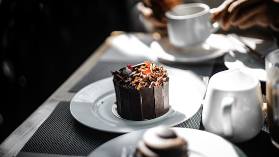 A dark chocolate dessert garnished with chocolate shavings and a small red fruit is placed on a white plate. Nearby, a person holds a white cup, suggesting a setting of dining or having a coffee. Other white crockery items are present on the table, which is covered with a textured, dark-colored mat, creating a contrasting background for the dessert.