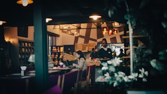 A cozy restaurant interior with dim lighting, featuring several tables where people are seated. A waiter is walking between the tables, and there are decorative elements like plants and shelves with displayed items on the walls. The ambiance is warm and inviting, with a gentle play of light and shadow.