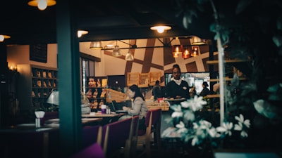A cozy restaurant interior with dim lighting, featuring several tables where people are seated. A waiter is walking between the tables, and there are decorative elements like plants and shelves with displayed items on the walls. The ambiance is warm and inviting, with a gentle play of light and shadow.