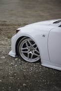 A close-up view of the front section of a white car with a sleek design. The focus is on the alloy wheel and the logo on the vehicle, which is parked on a gravel surface.
