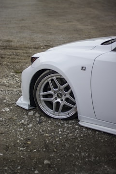 A close-up view of the front section of a white car with a sleek design. The focus is on the alloy wheel and the logo on the vehicle, which is parked on a gravel surface.