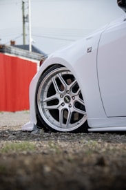 A close-up view of a car's front wheel featuring intricate silver alloy rims with a sporty design. The car is white and appears to be low to the ground, suggesting modifications. In the background, there is a red fence and part of a building with an overcast sky above.