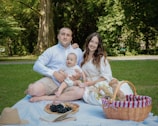 A cheerful family enjoys a picnic on a grassy area under large trees. They sit on a white blanket with a small child in the middle. A picnic basket, a straw hat, and various foods such as grapes and pastries are also visible. The scene reflects a serene outdoor environment.