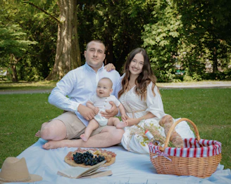A family enjoying a sunny afternoon picnic in a lush green park surrounded by pine trees.