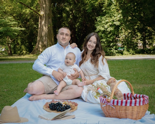 A cheerful family enjoys a picnic on a grassy area under large trees. They sit on a white blanket with a small child in the middle. A picnic basket, a straw hat, and various foods such as grapes and pastries are also visible. The scene reflects a serene outdoor environment.