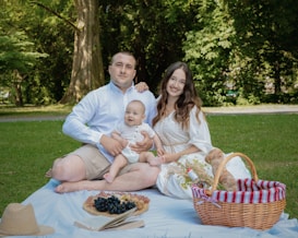 A cheerful family enjoys a picnic on a grassy area under large trees. They sit on a white blanket with a small child in the middle. A picnic basket, a straw hat, and various foods such as grapes and pastries are also visible. The scene reflects a serene outdoor environment.