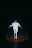 A person stands on a dimly lit stage under a spotlight, wearing a light-colored outfit and facing forward with arms slightly extended. A microphone is positioned in front of them, and stage lights are visible on the floor to either side.