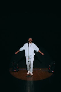 A person stands on a dimly lit stage under a spotlight, wearing a light-colored outfit and facing forward with arms slightly extended. A microphone is positioned in front of them, and stage lights are visible on the floor to either side.