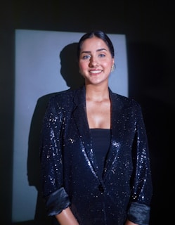 A person is wearing a dark, sequined blazer and is standing against a dark background with a lighter rectangular area behind them. The lighting creates a distinct shadow on the wall. The person's hair is neatly styled, and they are smiling at the camera.