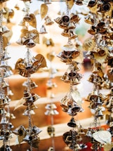 Strings of small, shiny brass bells and decorative leaf-shaped ornaments hang closely together. The background is softly blurred, highlighting the intricate details and reflective surfaces of the bells and ornaments.
