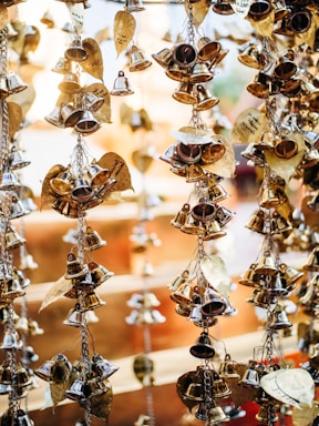 Strings of small, shiny brass bells and decorative leaf-shaped ornaments hang closely together. The background is softly blurred, highlighting the intricate details and reflective surfaces of the bells and ornaments.