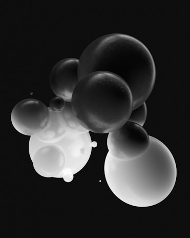 An arrangement of various gray spheres floating in a dark, possibly black environment. The spheres vary in size and appear to have a matte surface, with subtle reflections and shading that give them a three-dimensional appearance.
