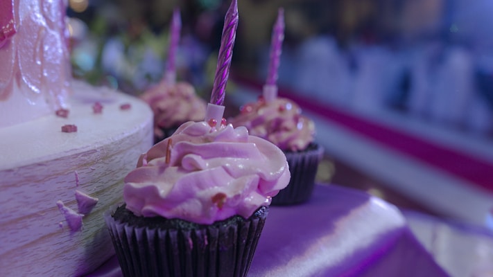 Cupcakes with swirled pink frosting, each topped with a pink candle, are arranged on a table with a purple cloth. A partially visible cake is in the background, reflecting a festive mood.
