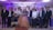 A group of people, mostly young men, are gathered indoors dressed in formal attire, centered around a young woman wearing an elegant peach-colored gown and holding a bouquet of flowers. The backdrop is ornately decorated with purple and white floral arrangements and atmospheric lighting.