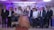 A group of people, mostly young men, are gathered indoors dressed in formal attire, centered around a young woman wearing an elegant peach-colored gown and holding a bouquet of flowers. The backdrop is ornately decorated with purple and white floral arrangements and atmospheric lighting.
