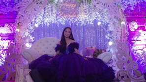 A woman wearing a beautiful dark purple gown is seated on an elegant white chair. She is surrounded by an elaborate decorative setup featuring white floral arrangements and butterfly motifs. The backdrop is adorned with sparkling elements and soft lighting, with the words 'Happy Birthday' illuminated above her. Several floral gifts and arrangements are also visible around her.