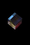 A transparent cube made of glass or crystal, resting against a black background. Light refracts through the cube, creating color gradients of red, yellow, blue, and green. The cube has a polished surface that reflects light, giving it a shiny appearance.