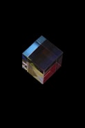 A transparent cube made of glass or crystal, resting against a black background. Light refracts through the cube, creating color gradients of red, yellow, blue, and green. The cube has a polished surface that reflects light, giving it a shiny appearance.