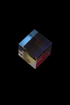 A transparent cube made of glass or crystal, resting against a black background. Light refracts through the cube, creating color gradients of red, yellow, blue, and green. The cube has a polished surface that reflects light, giving it a shiny appearance.
