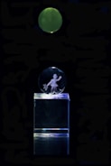 A clear acrylic glass display featuring an engraving of a person holding an item, mounted on a transparent rectangular base. Above the display, there is a green, glowing circular object against a completely dark background.