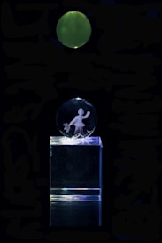 A clear acrylic glass display featuring an engraving of a person holding an item, mounted on a transparent rectangular base. Above the display, there is a green, glowing circular object against a completely dark background.