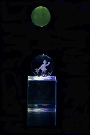A clear acrylic glass display featuring an engraving of a person holding an item, mounted on a transparent rectangular base. Above the display, there is a green, glowing circular object against a completely dark background.