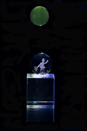 A clear acrylic glass display featuring an engraving of a person holding an item, mounted on a transparent rectangular base. Above the display, there is a green, glowing circular object against a completely dark background.
